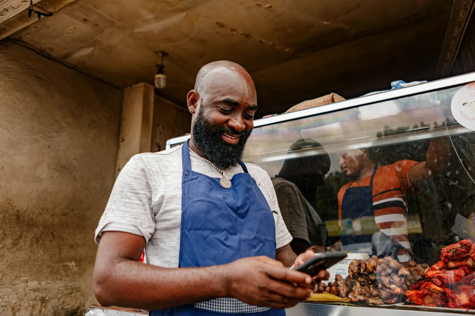 Food stall owner in a blue apron smiling down at his phone behind a glass counter of grilled meat
