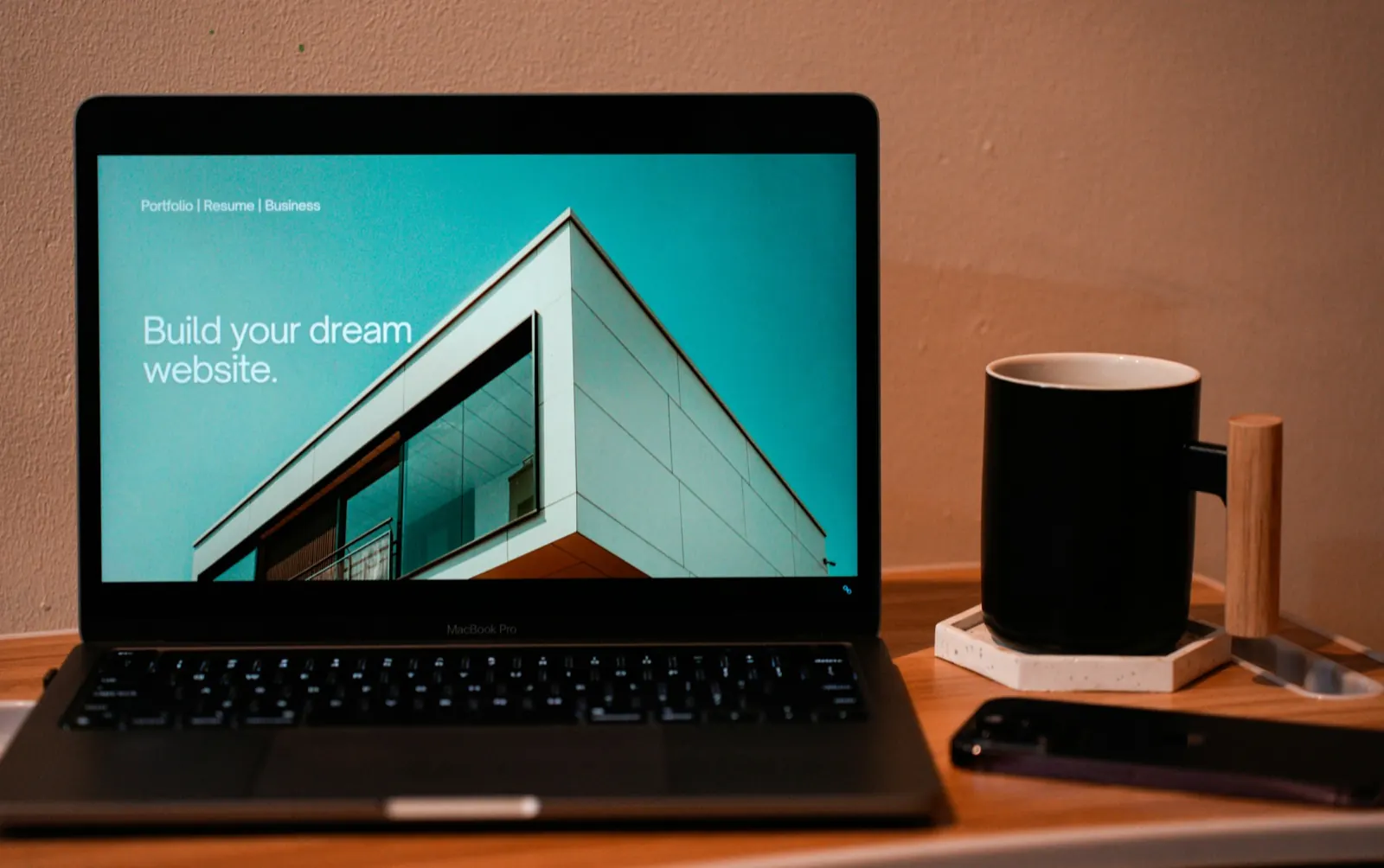 Laptop on a wooden desk showing a teal website page with a modern building and the headline Build your dream website, beside a black mug and a phone